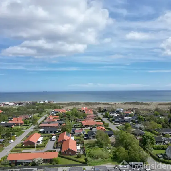 Boligområde med røde tage strækker sig mod horisonten, set fra oven. Baggrunden domineres af havet og en blå himmel med spredte skyer.