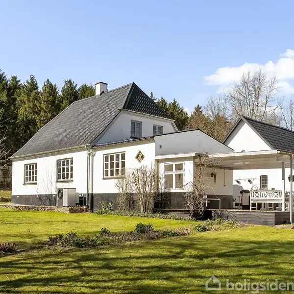 Hus i traditionel stil med sort tag og hvide vægge, står på en grøn græsplæne. Omkring er der skov og blå himmel. Terrasse med overdækning ses ved siden af huset.