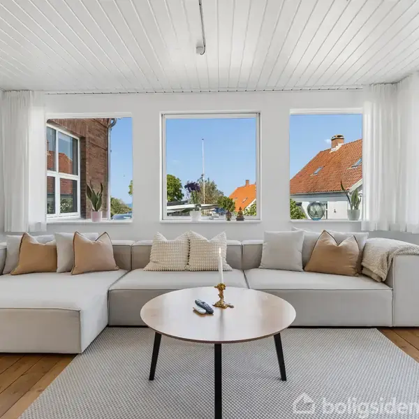 En stor, lys sofa står i en stue med trægulv. Et rundt sofabord foran sofaen holder en lysestage. Store vinduer giver udsigt til nabohuse og en blå himmel.