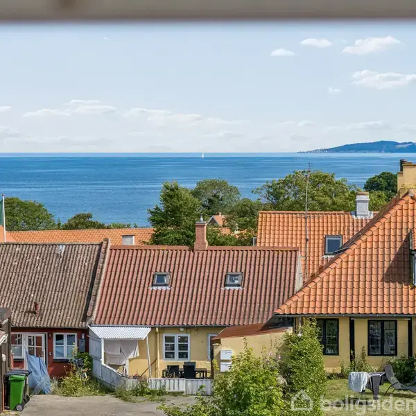 Et hvidmalet murstenshus med rødt tag står stille på en rolig villavej. Baggrunden viser flere huse og en blå himmel med få skyer.