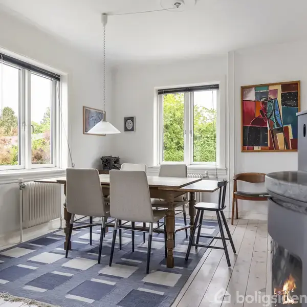 Et spisebord står centralt med seks stole omkring det. Bordet er placeret på et tæppe i et lyst rum med store vinduer og kunst på væggene. En brændeovn ses til højre.