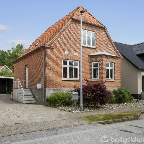 Rødt murstenshus med skarpt, rødligt tegltag, har inskriptionen “Elisabog” på facaden. Det står ved en gade med fortov, omgivet af andre huse og grøn beplantning foran.