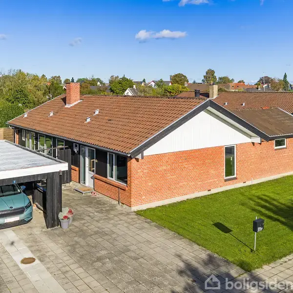 En rød murstensvilla med brunt tag står ved siden af en carport, der indeholder en bil. Omgivet af en velplejet grøn plæne i et boligområde.
