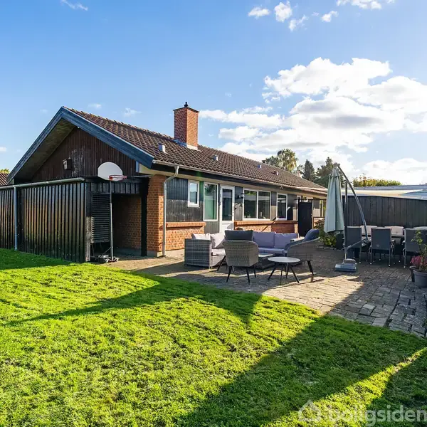 Et murstenshus med store vinduer og skorsten står i solen. En møbleret terrasse med parasol og stole ligger ved siden af. Græsplæne omgiver huset.
