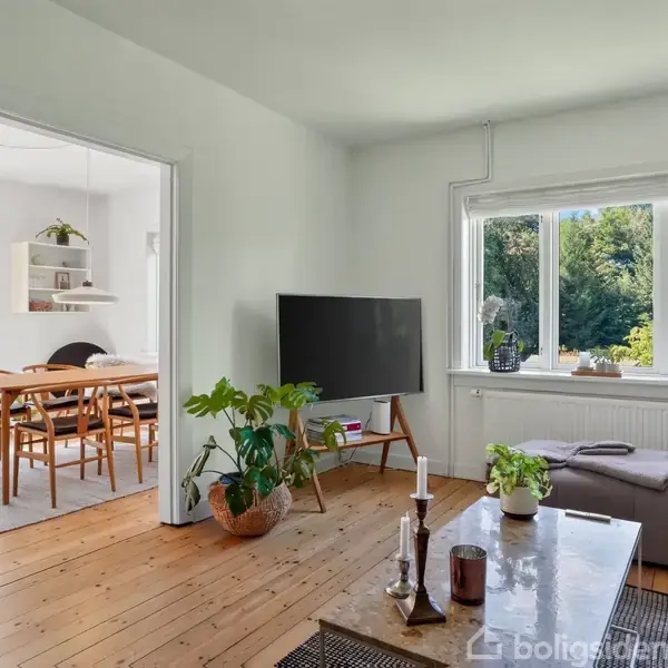 Stue med grå sofa og TV på stander, står ved siden af vindue med udsigt til have. Åben passage til spisestue med træbord og designerstole. Planter og pynt genstande dekorerer rummet.