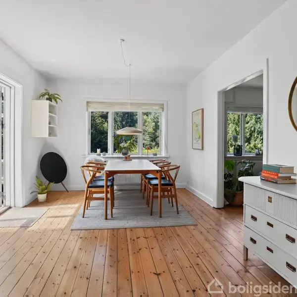 En rektangulær spisebord med seks stole står midt i et lyst spisestue med trægulv. Vægge er hvide, og der er store vinduer med udsigt til en grøn have.