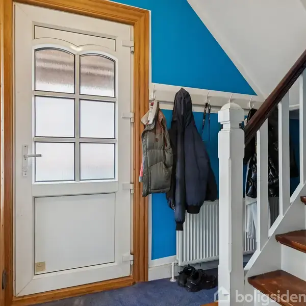 Dør står stille i en entré med blå vægge og træramme. Overtøj hænger på knager ved siden af en trappe med hvide balustre og trætrin.