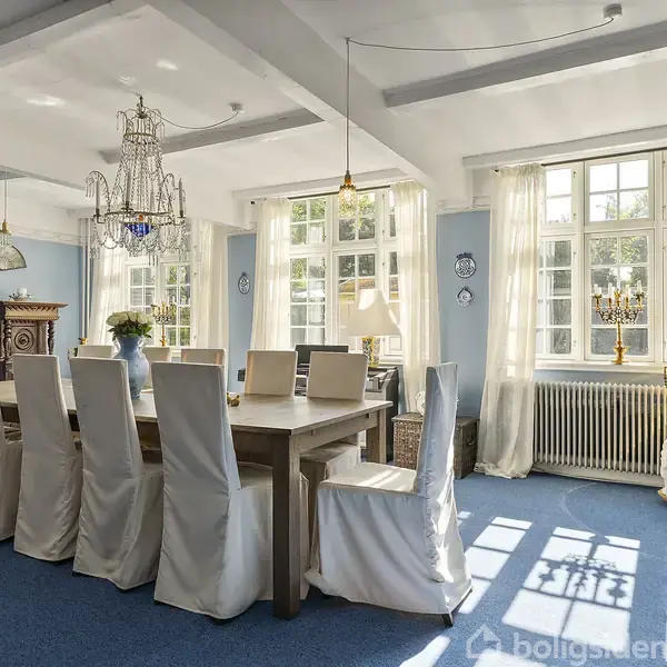 Langt spisebord med ti stole dækket med hvide stofovertræk. Store vinduer med gardiner tillader dagslys ind i rummet. Elegant lysekrone hænger fra loftet. Vægge er blå.