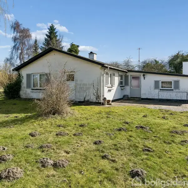 Hus står stille i en have med græs og spredte buske. Omkring er der træer og en klar blå himmel i baggrunden.