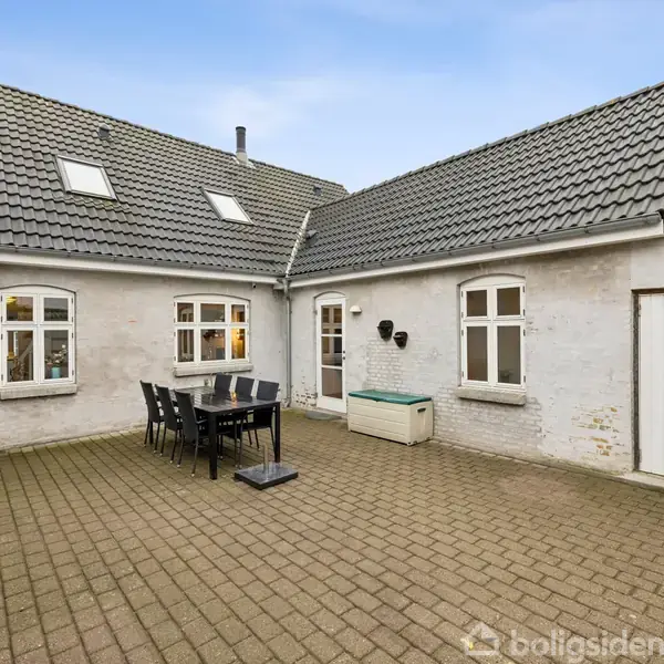 En gårdhave med flisebelægning indeholder et spisebord med stole. Omkring gårdhaven er et hus med hvide vægge og sorte tagsten. Et skur eller rum med hvid dør ses til højre.