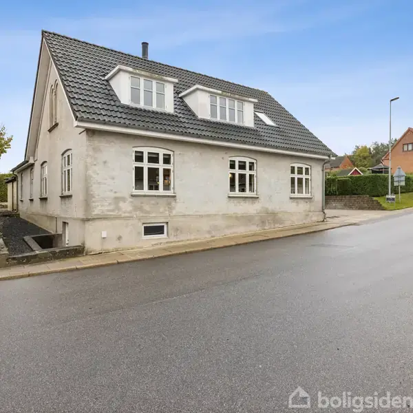 Huset med hvid facade og gråt tag står på en stille vej. Det har flere vinduer og ligger i et beboelseskvarter med græs og nabohuse omkring.