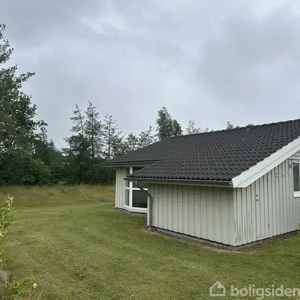 Et hvidt træhus står stille på en velplejet græsplæne omgivet af træer. Taget er mørkt, og en rød bil er delvist synlig på højre side.