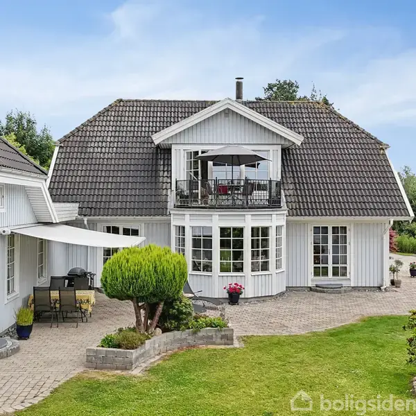 Et hvidt træhus med mørkt tag står på en velplejet grund. En lille terrasse med møbler befinder sig under en markise, omkranset af grønne buske og træer.