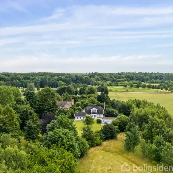 Et hvidt hus med mørkt tag står stille i en frodig, grøn skov. Omgivelserne udgøres af åbne marker og en blå himmel.
