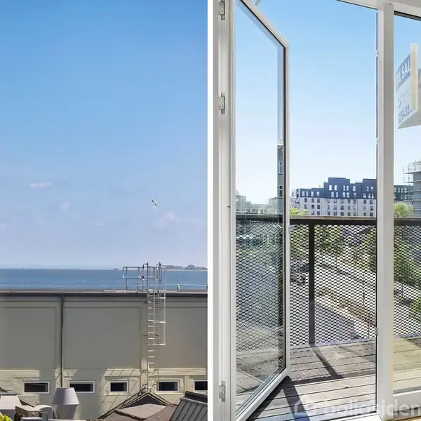 Åben terrassedør fører til en balkon med udsigt over en bylandskab og havet i baggrunden. Nogle bygninger og træer ses under en klar blå himmel.