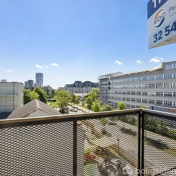 En balkon med netgelænder, der vender ud mod en byudsigt. Der ses flere bygninger, træer og en klar himmel. Skiltet viser "EDC", "Poul Du...," "32 54 32 54" i øverste højre hjørne.