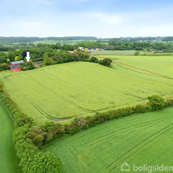 Luftfoto viser en stor grøn mark indrammet af træer, med en rød bygning peget ud af en hvid pil. Landskabet er bakket med skove og marker i baggrunden.