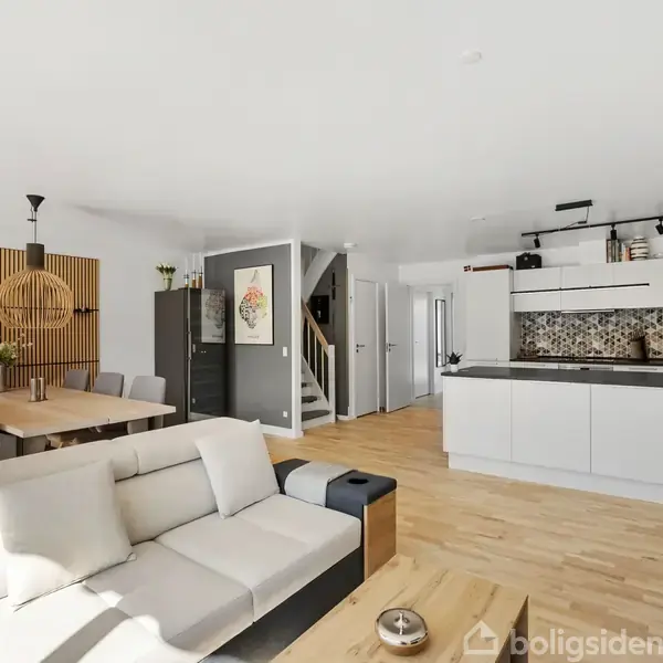 Sofa i en åben stue; placeret mod et køkken med hvide skabe og sort bordplade. Der er et spisebord med stole og vægudsmykning af træpaneler.