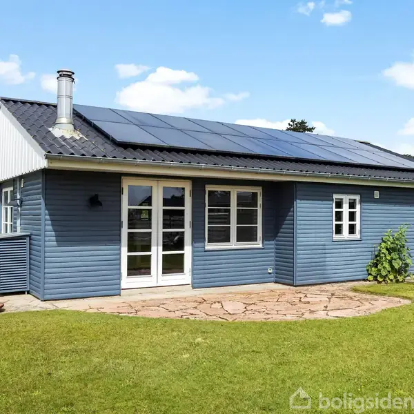 Blåt hus med solpaneler på taget står stille i en velplejet have med stenfliser og grøn græsplæne, under en lys blå himmel med spredte skyer.