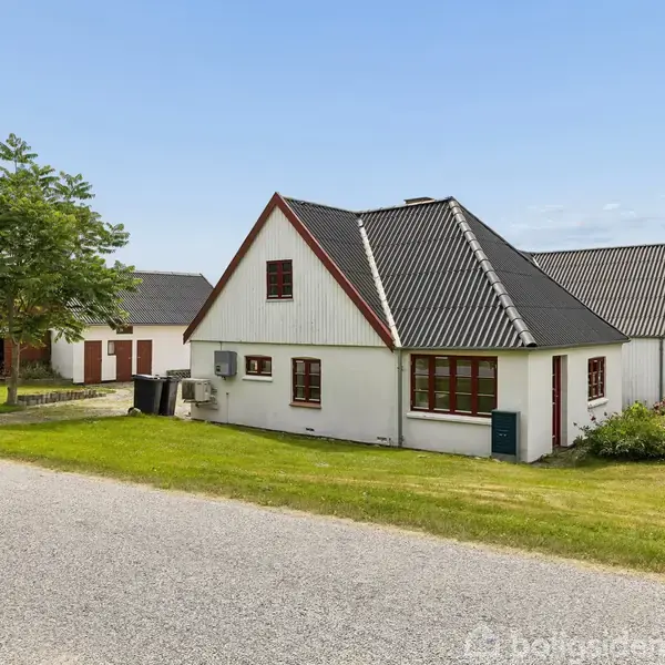 Et hvidt hus med rødt tag står på en grøn græsplæne ved en vej. Ved siden af ses en mindre bygning og et stort træ.