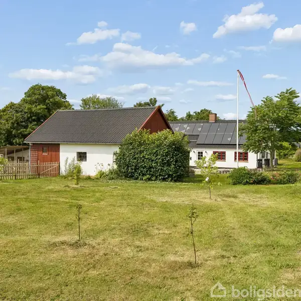 Et hvidt hus med rødt tag ligger ved en vej, omgivet af en grøn have og træer. Solpaneler er synlige på taget.