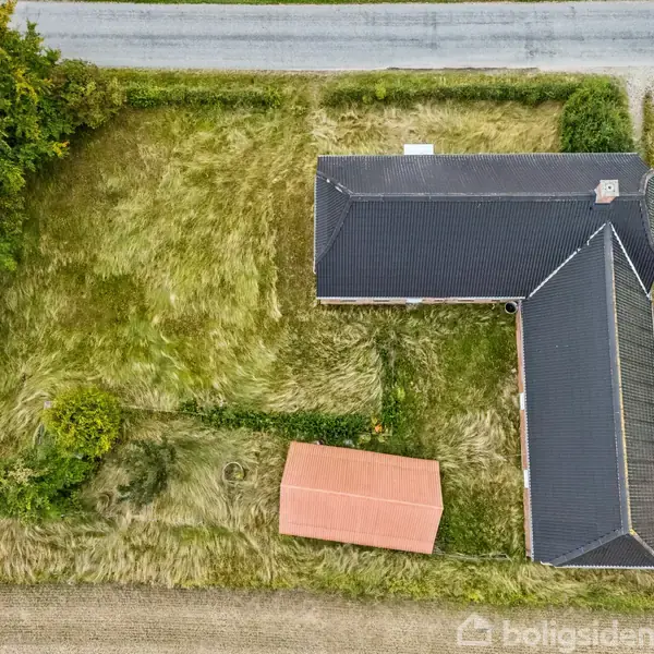 Hus med sort tag i L-form, set fra oven, beliggende på en græsplæne ved siden af en vej. To røde udhuse ses tæt på huset. Omkranses af grønt landskab.