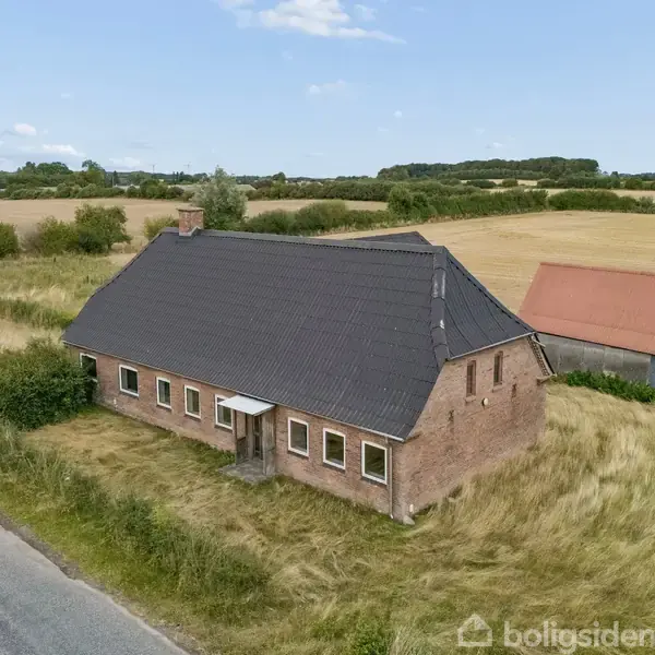 En ældre, murstensbygget landejendom med sort tag står stille blandt høje græsser ved en landevej. I baggrunden ses en rød lade og åbne marker.