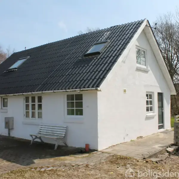 Et hvidt hus med sort tag har ovenlysvinduer, stående i en have med bladløse træer. En bænk står foran huset på flisebelægningen.