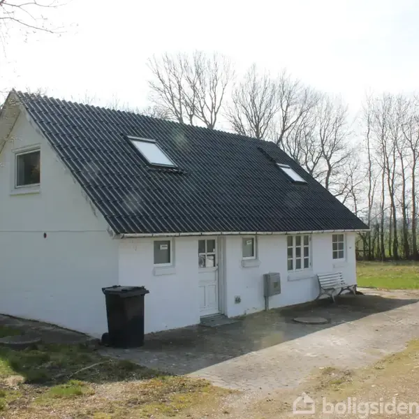 Et hvidt hus med mørkt tag står i en grøn have. Der er to døre og tre vinduer på facaden. Baggrunden har nøgne træer og græsareal.