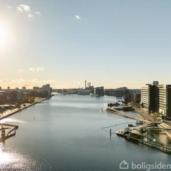 Sollys skinner over en bred flod omgivet af moderne kontorbygninger. Gaderne og kajområdet ved floden er stille, hvilket skaber en fredelig byatmosfære.