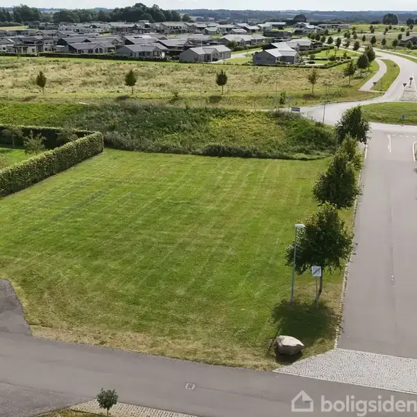 En stor, grøn græsplæne ligger stille mellem asfaltveje omkranset af buskads og hækker i et boligområde med mange huse i baggrunden. En bil ses på hjørnet af vejen.