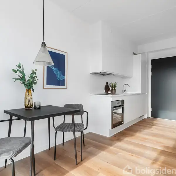 Et lille køkken med hvidt køkkenudstyr og en indbygget ovn ved siden af et lille spisebord med to stole. En vase med grønne planter står på bordet.