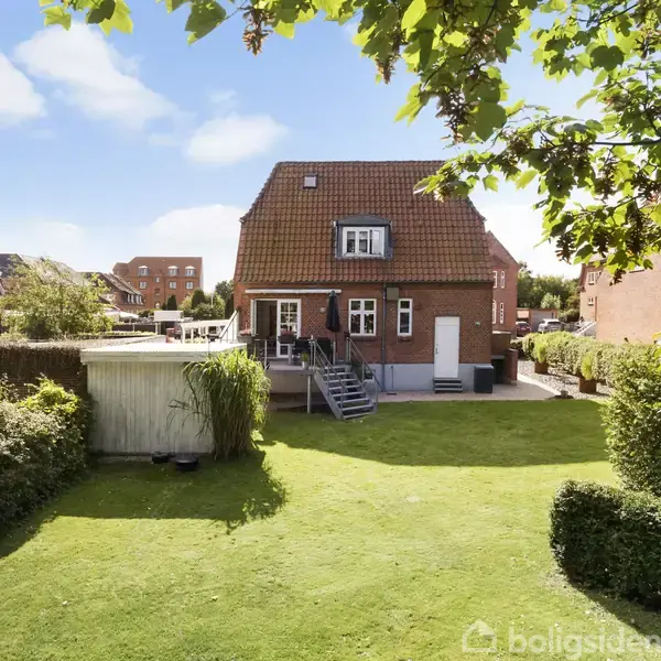 Rødt murstenshus med kvist og træterrasse, omgivet af grøn have med græsplæne. Træer og buske rammer huset ind i et villakvarter.