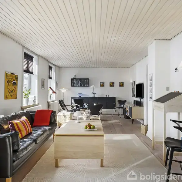 En stue med en sort lædersofa, sofabord og tv. Væggene er hvide med kunst. Store vinduer lader lys strømme ind. Indretningen er moderne og lys.