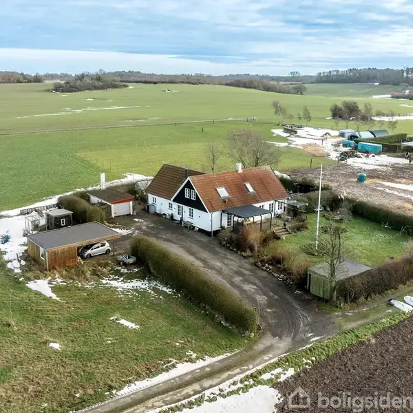 Et hvidt hus med rødt tag står blandt sne og græsarealer. Der er et skur ved siden af. Omgivet af marker og en grusvej.