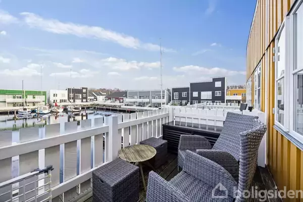 Fletstole og bord står stille på en altan med udsigt over en havn med moderne bygninger i forskellige farver.