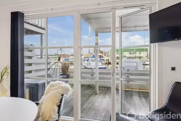 Et spisebord med en stol med pelsbetræk står indenfor, mens glasdøre åbner ud til en terrasse med udsigt over en havn, hvor både er fortøjet.