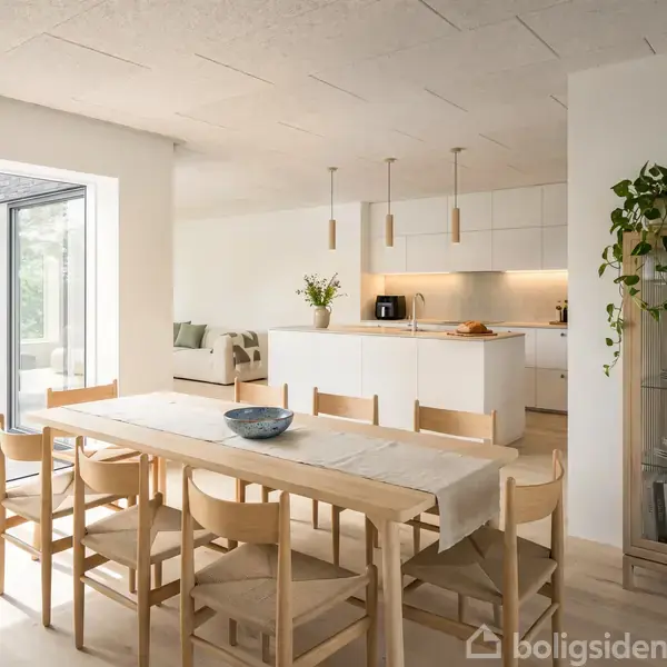 En spisestue med træbord og stole, set mod et moderne hvidt køkken. Der er planter og en vitrineskab. Store vinduer til venstre giver naturligt lysindfald.