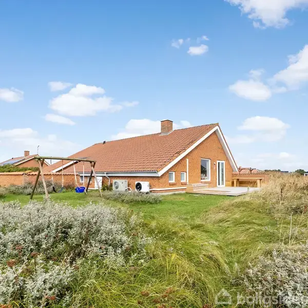 Et hus med rødt tegltag står i en grønt, græsbevokset naturområde. En gyngestativ er placeret ved siden af huset under en blå himmel med få skyer.