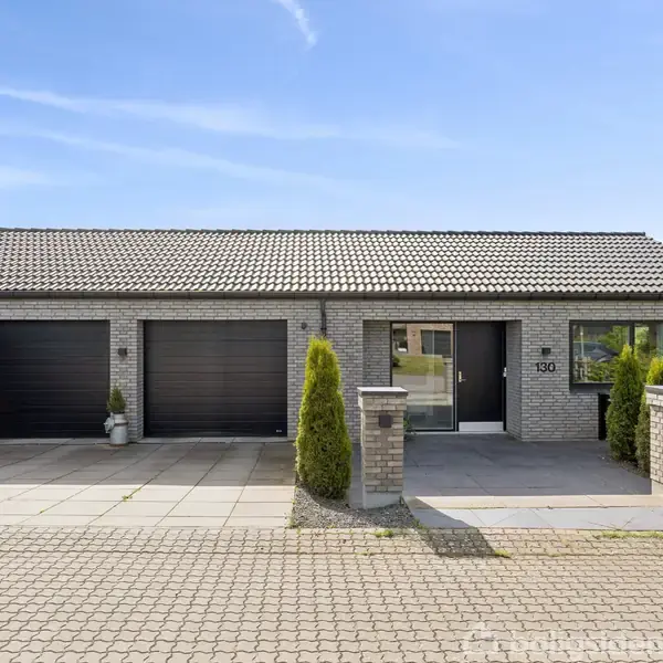 En moderne bungalow med to garageporte og en enkelt indgangsdør, beliggende i et forstadsområde. Foran huset står et par grønne buske, og der er husnummer "130" ved døren.