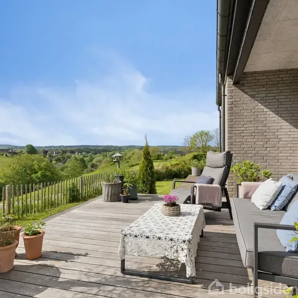 Terrasse med grå sofa og lænestole, hvor planter på borde og i krukker dekorerer området. Solbeskinnet landskab strækker sig ud i baggrunden, med frodige grønne marker og en blå himmel.