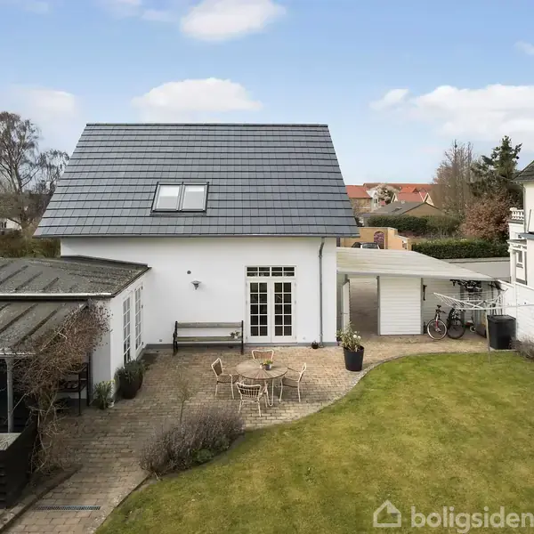Et hvidt hus med skråt tag og terrassedør står i en have med græsplæne og flisebelagt terrasse. Til venstre ses et sort havehus og cykler under et halvtag.