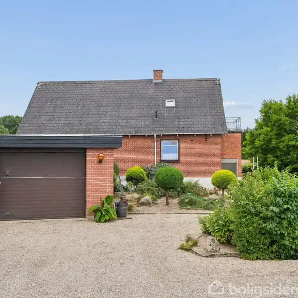 Rødt murstenshus med sadeltag, står for enden af en grusindkørsel, omgivet af grønne buske og en garage til venstre. Klar blå himmel i baggrunden.
