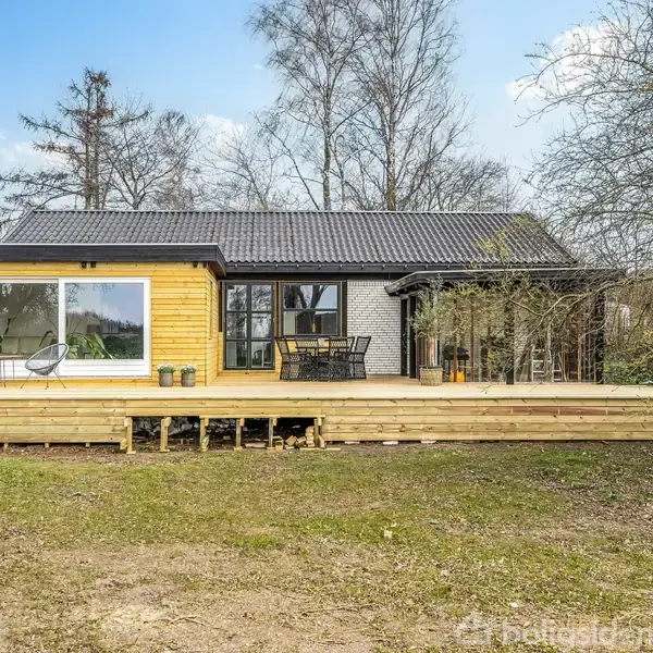 Et moderne sommerhus med store vinduer og en træterrasse står i en haveomgivelser med græsplæne og træer. Terrasse med hvide havemøbler. Ingen tekst til stede i billedet.