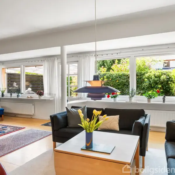 Stue med sorte sofaer og et sofabord med en vase med blomster. Store vinduer bagved giver udsigt til en grøn have. En lampe hænger fra loftet, og der er tæpper på gulvet.