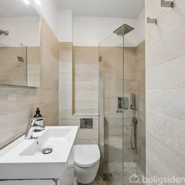 Hvide vask og toilet står i et moderne badeværelse med beige fliser. Bruseniche med glasvæg er til højre. Et spejl hænger over vasken. Ingen tekst i billedet.