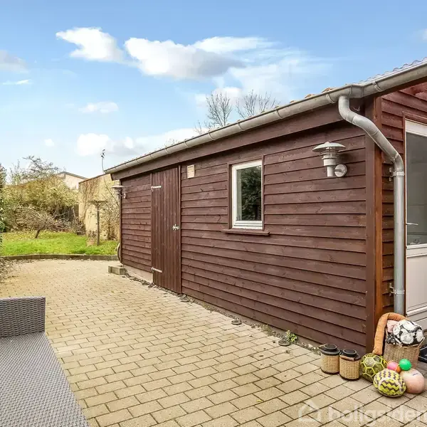 En træbeklædt bygning med et lille vindue og en hvid dør fører til en terrasse med grå fliser i en have. Ved døren står flere farvede potter.