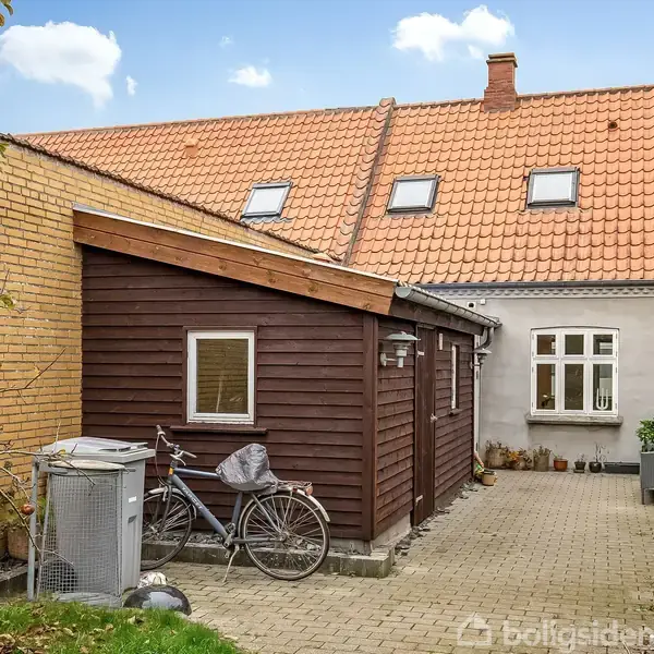 En cykel står parkeret ved en brun træskurvæg; i baggrunden ses et hus med rødt tegltag og flere vinduer. Omgivende murstensvægge og brostensbelægning skaber en hyggelig gårdhave.