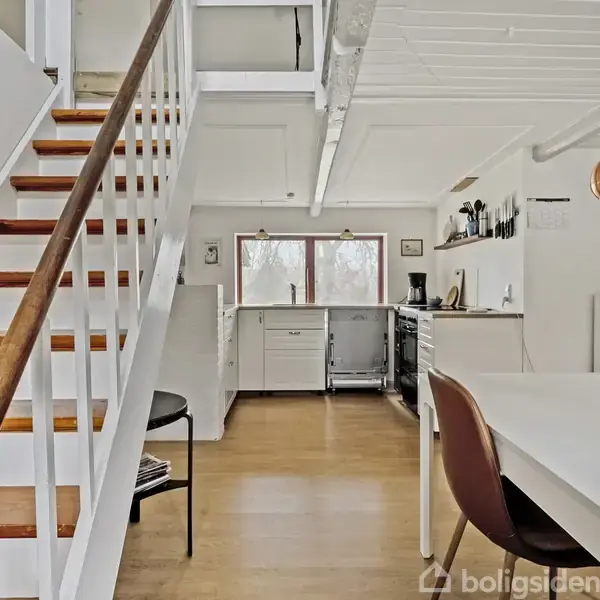 Trappen med trætrin fører op i et lyst køkken med hvide skabe. Der er en hvid bordplade, brune stole og en kobberfarvet lampe. Store vinduer giver naturligt lys.