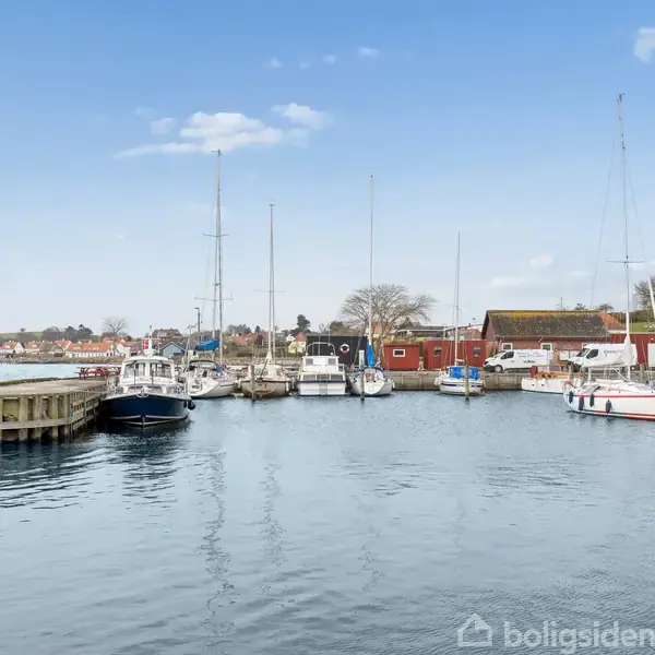 Både ligger fortøjet ved en rolig havn; røde bygninger ses i baggrunden under en klar himmel med spredte skyer.